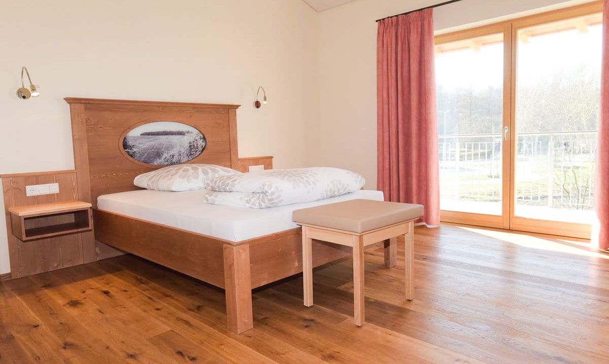 A well-appointed room features a spacious bed with a wooden headboard and decorative mirror. Natural light floods the space through glass doors leading to a balcony, complemented by light-colored curtains. A simple wooden bench near the bed and a small nightstand enhance the functionality of the room.