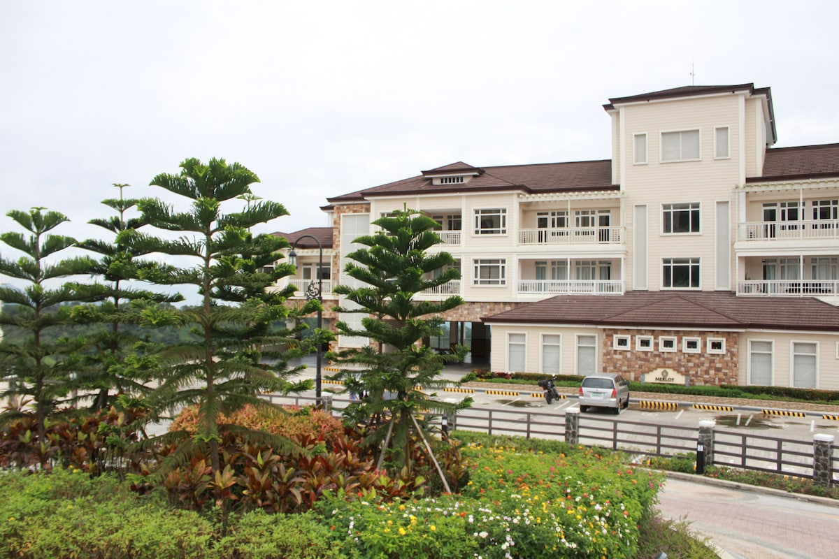 The exterior of Merlot Residences is displayed with neatly arranged greenery and colorful flower beds in the foreground. The three-story building features balconies and large windows, blending with the natural surroundings and offering a serene atmosphere.