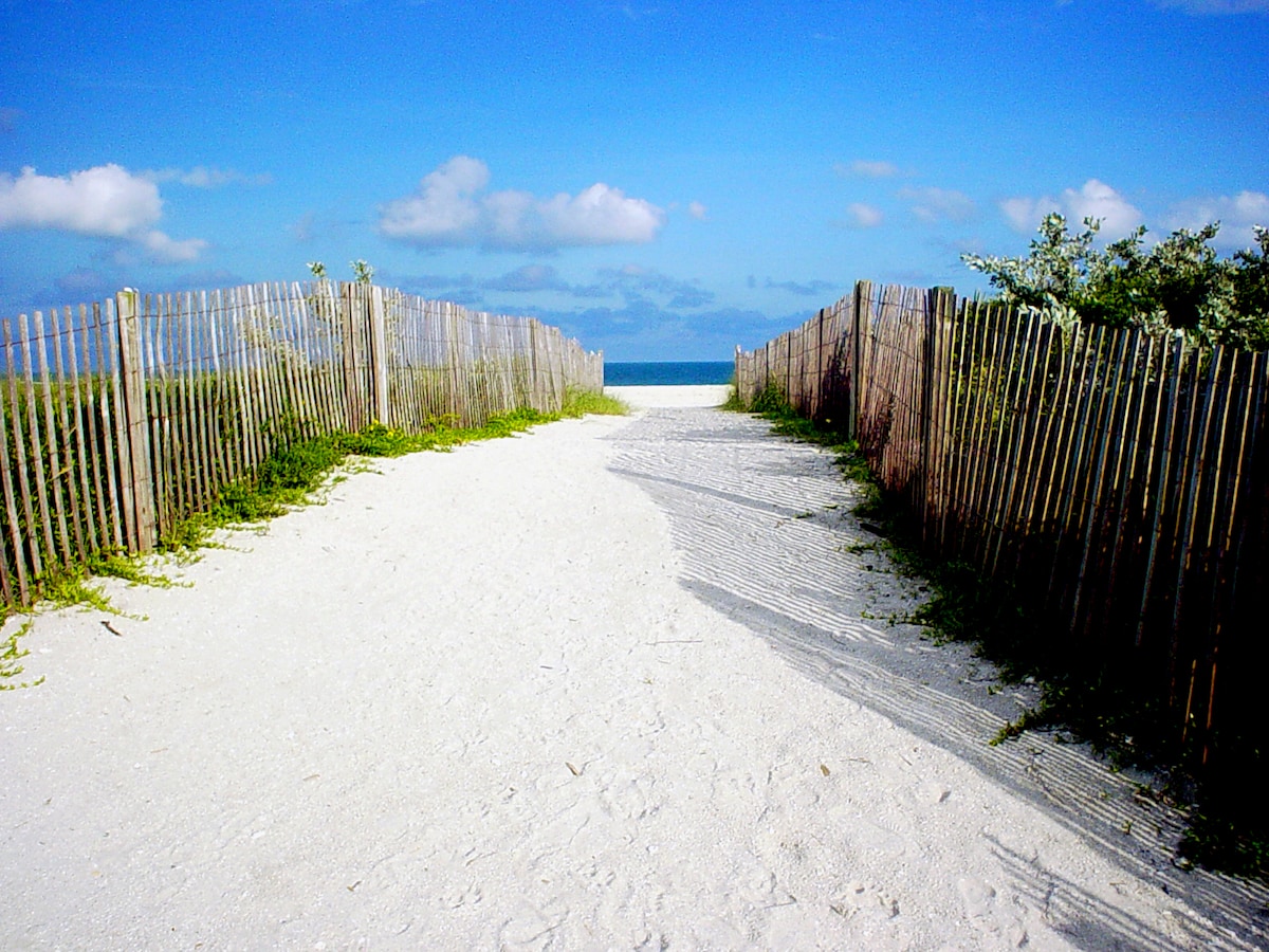 A sandy pathway leads through two wooden fences towards the beach, framed by natural vegetation. The bright blue sky and scattered clouds create a serene backdrop, inviting guests to enjoy the coastal atmosphere just a short walk away.