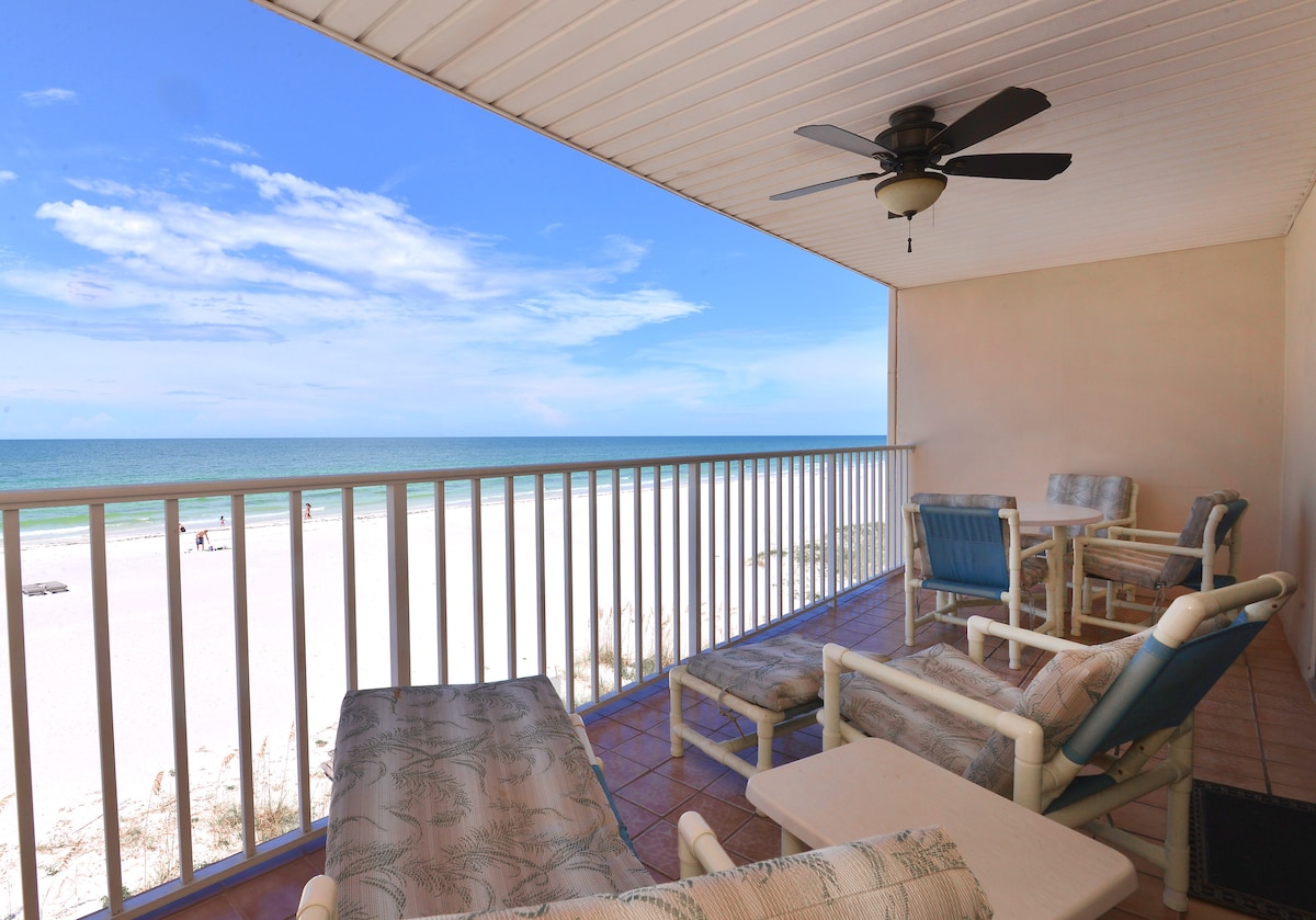 A private balcony with a direct view of the Gulf of Mexico features a round table with four chairs and a lounge chair. A ceiling fan is installed overhead, and the soothing colors of the sea are visible beyond the railing.