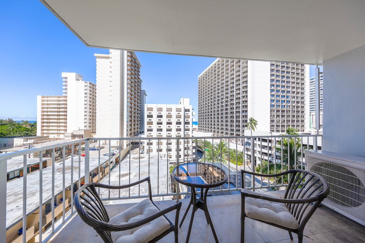 A private terrace features a small round table and two comfortable chairs, surrounded by views of nearby buildings and palm trees. Clear blue skies are visible above, enhancing the open atmosphere.