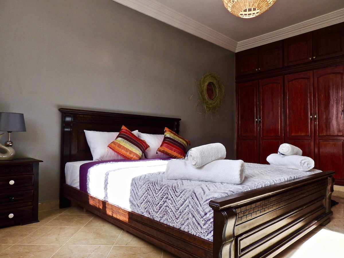 A spacious double room is furnished with a large wooden bed topped with white linens and decorative pillows. Soft towels are neatly arranged on the bed. A dark wooden nightstand with a lamp is placed beside the bed, and ample storage is provided by the fitted wardrobes in the background.