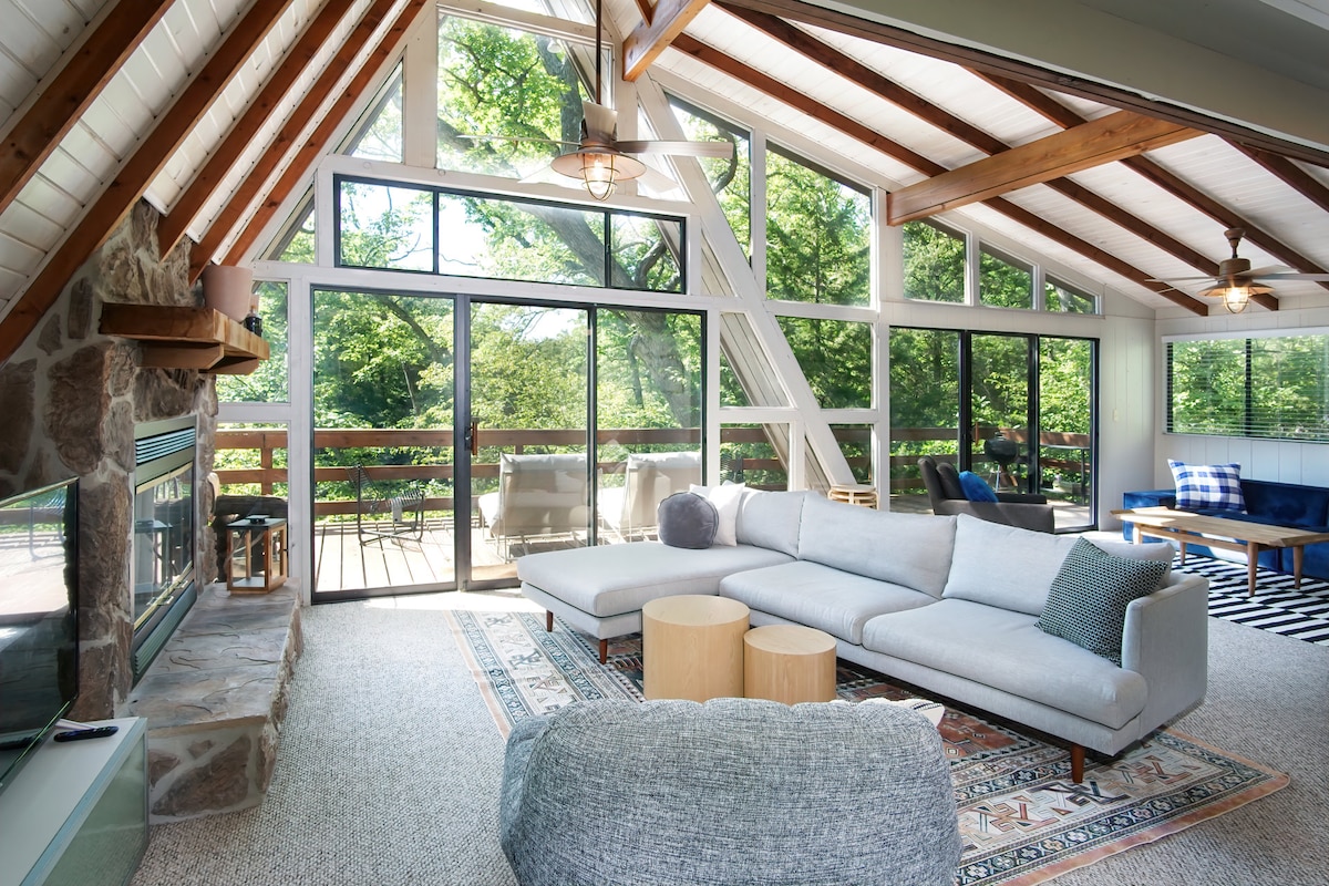 The living room features a large sectional sofa placed on a patterned area rug, adjacent to two cozy accent chairs. Expansive windows provide a view of the surrounding greenery and allow natural light to fill the space, while a stone fireplace adds a rustic touch.