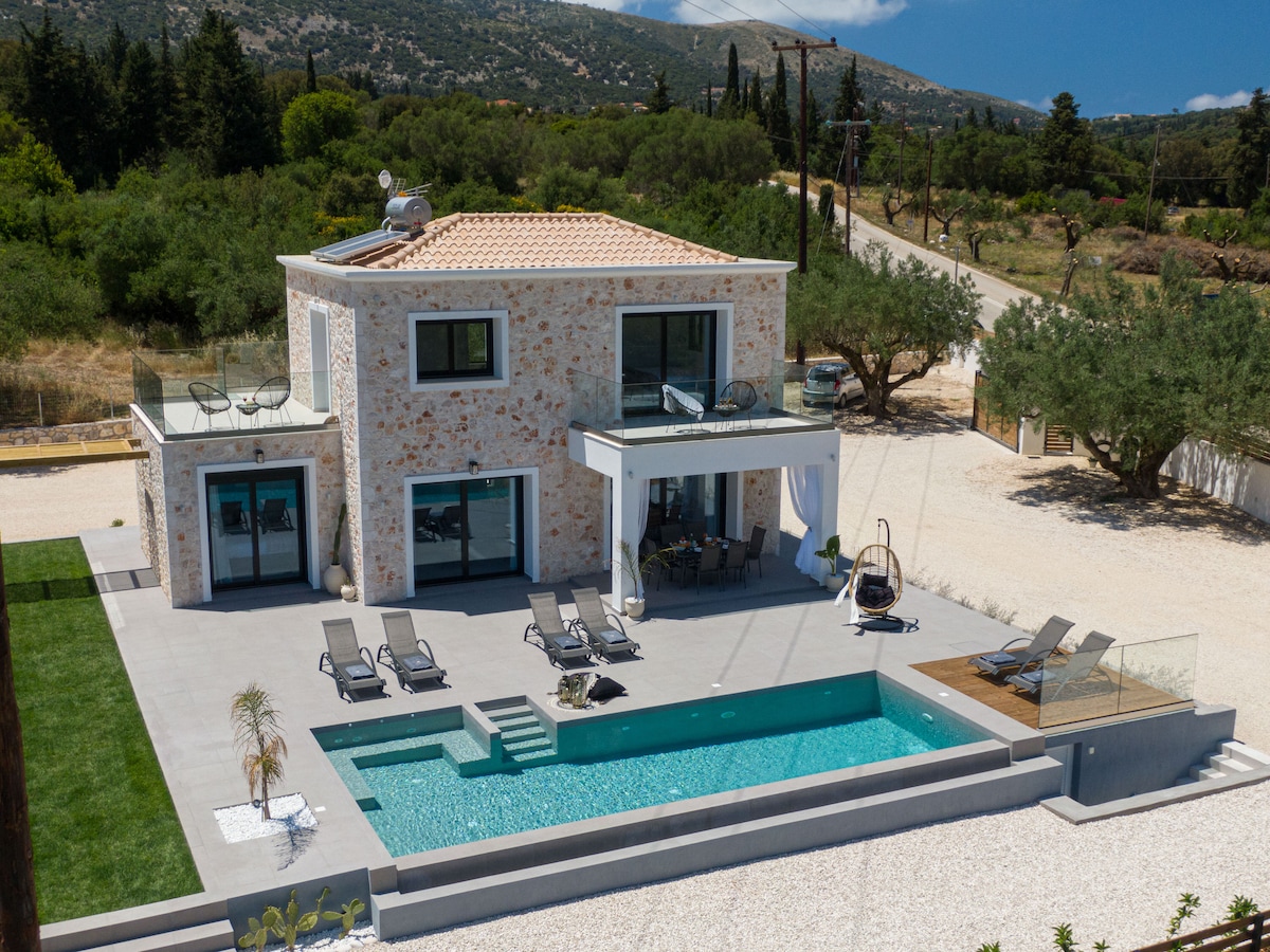 A modern villa is surrounded by landscaped gardens and features a private infinity swimming pool, complemented by sunbeds and outdoor seating. The spacious terrace is designed for relaxation, with a swing chair and mountain views in the background, reflecting a serene outdoor living environment.