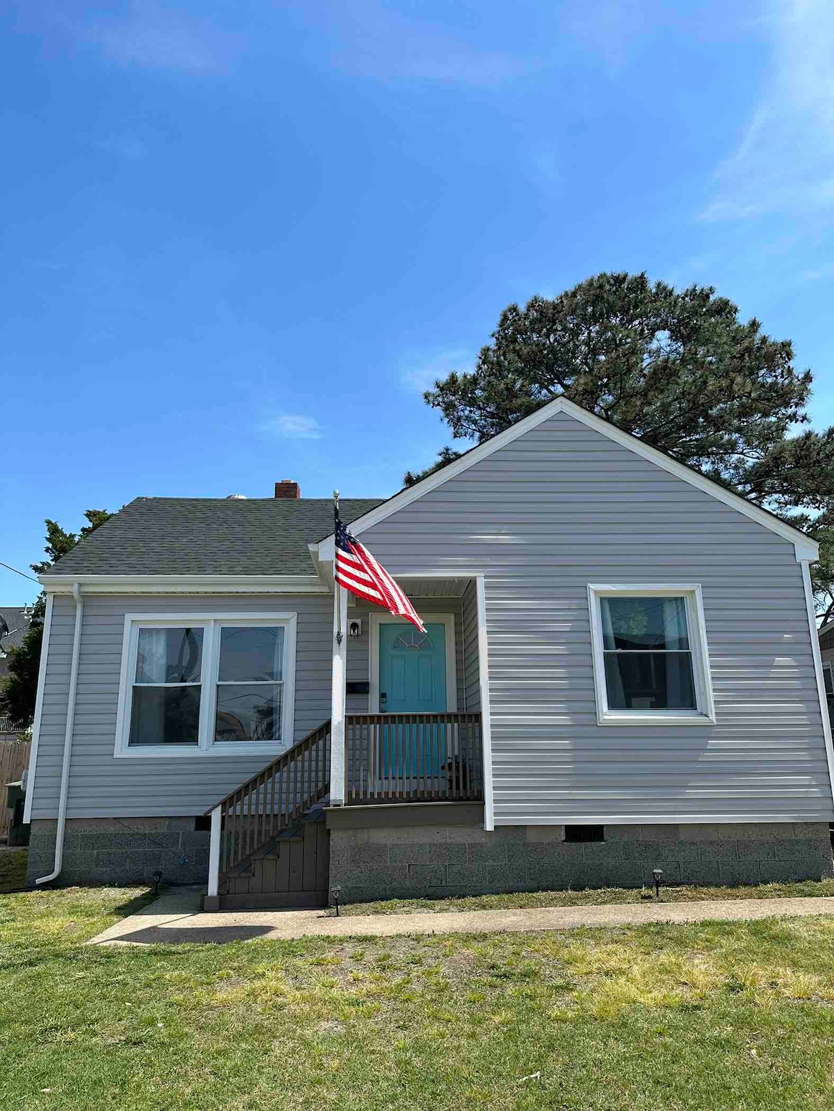 A single-story beach house is shown with light gray siding and a sloping roof. An American flag proudly waves from the porch. Large windows provide natural light to the interior, and a small set of stairs leads to the entrance, flanked by a neat lawn.