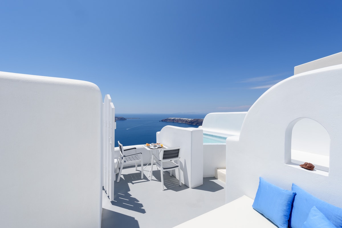 An outdoor terrace features white walls and a light grey floor, providing a spacious area equipped with a small dining table and chairs. A bright blue cushion adds a touch of color. The view includes the sea and distant islands under a clear blue sky.