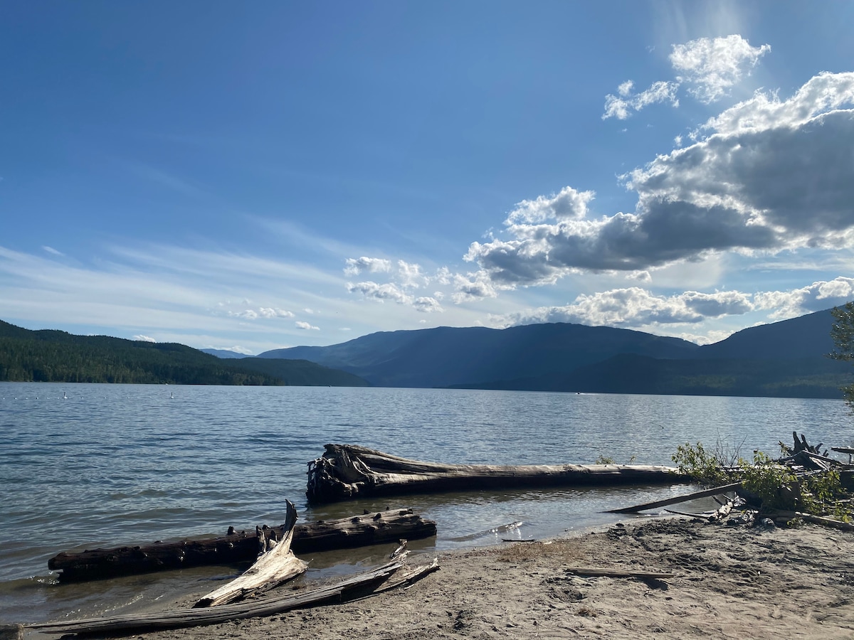 A serene lakeside view features calm waters reflecting the sky, with scattered clouds overhead. Fallen logs rest on the sandy shoreline, while mountains rise in the background, creating a tranquil natural setting.