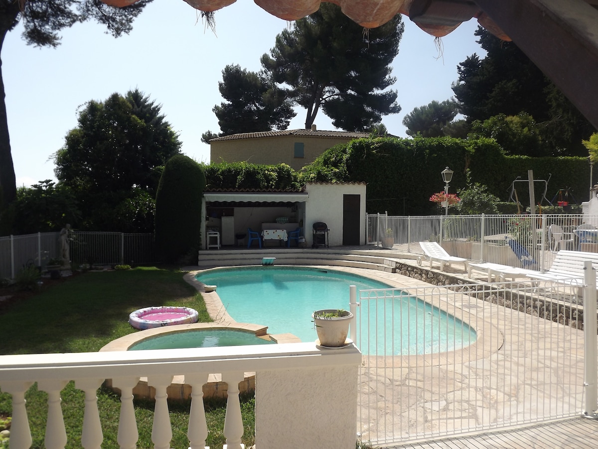 A well-maintained swimming pool is surrounded by a spacious deck and secured by a fence. Lush greenery and tall trees are visible in the background, offering shade and privacy. A shaded seating area is positioned beside the pool, creating a relaxing environment.