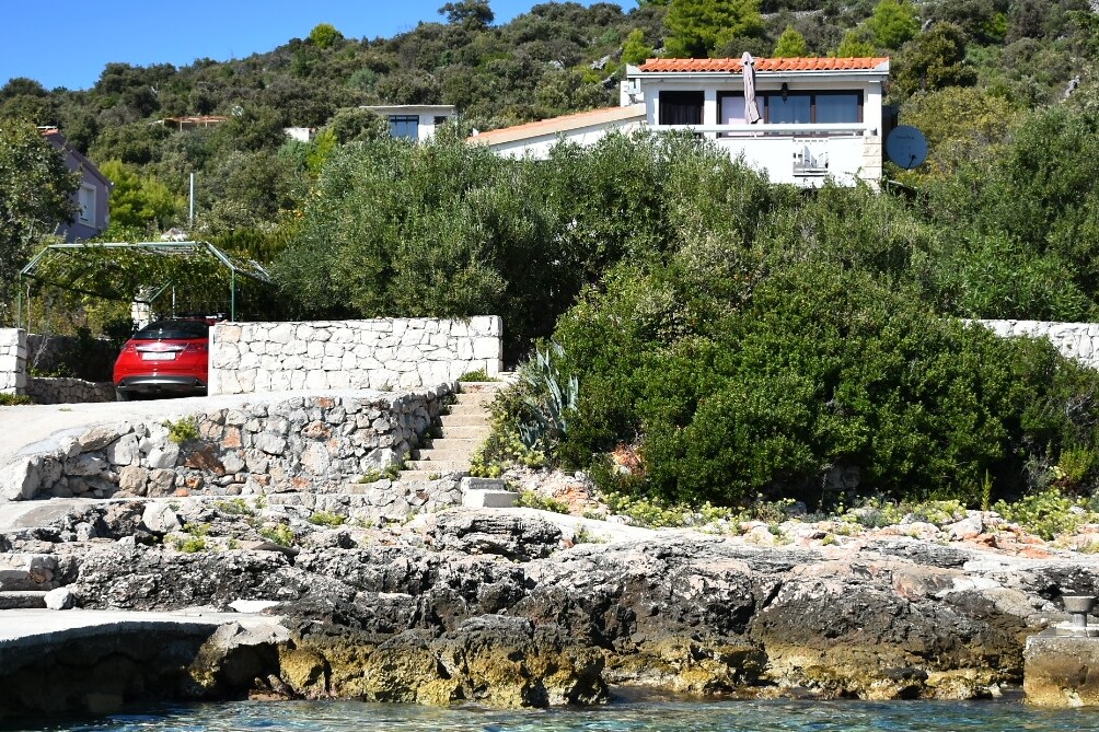 The house is set atop a lush, green hillside, surrounded by thriving plants. A stone pathway leads up to the entrance, with a carport visible at the base. Clear blue waters gently lap at the rocky shoreline in the foreground.
