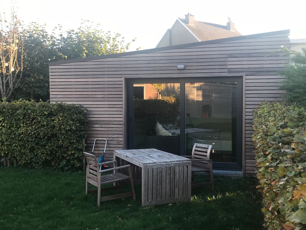 The exterior of a modern tiny house is presented, featuring wooden siding and large glass doors. A small outdoor table and two chairs are positioned on a grassy area, surrounded by neatly trimmed hedges and shrubbery.