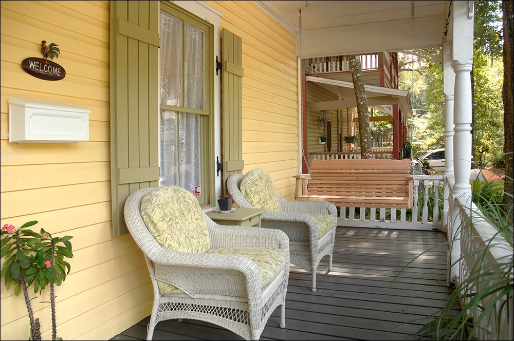 A peaceful front porch features two wicker chairs with floral cushions, positioned on a wooden floor. A small side table rests between the chairs, and a welcoming sign is mounted near the entrance. A porch swing is visible in the background, surrounded by greenery.