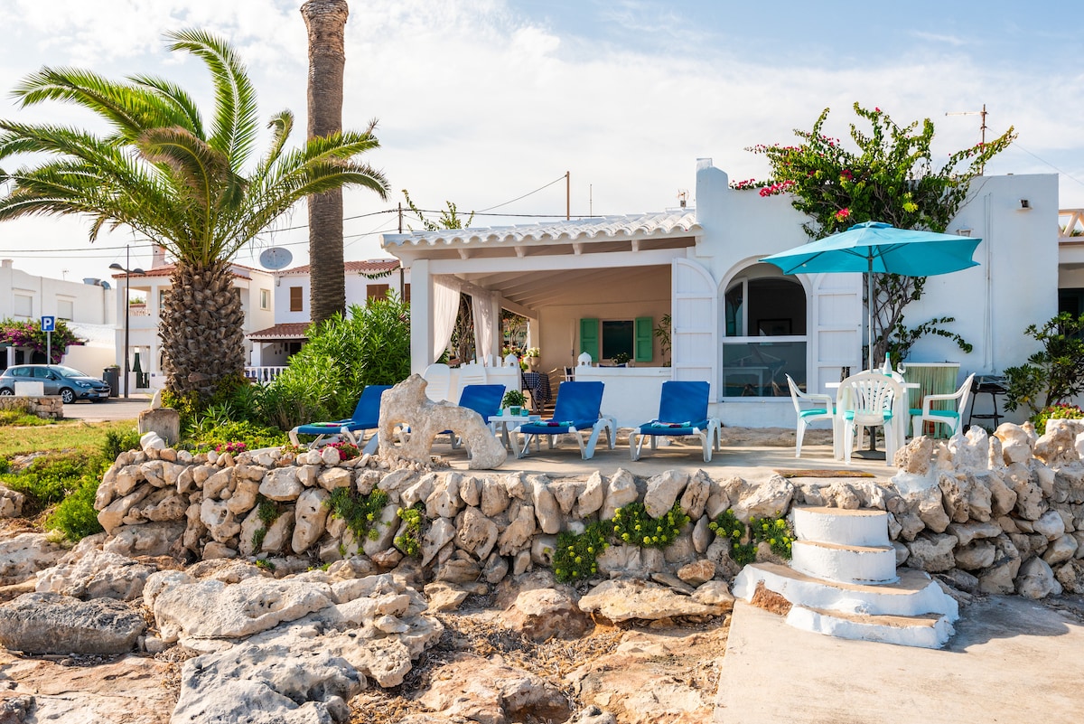 A terrace is adorned with lounge chairs and a small table, surrounded by stone walls. A bright blue umbrella provides shade, while palm trees and lush greenery are visible in the background. The setting offers direct views of the coastline, enhancing the outdoor experience.