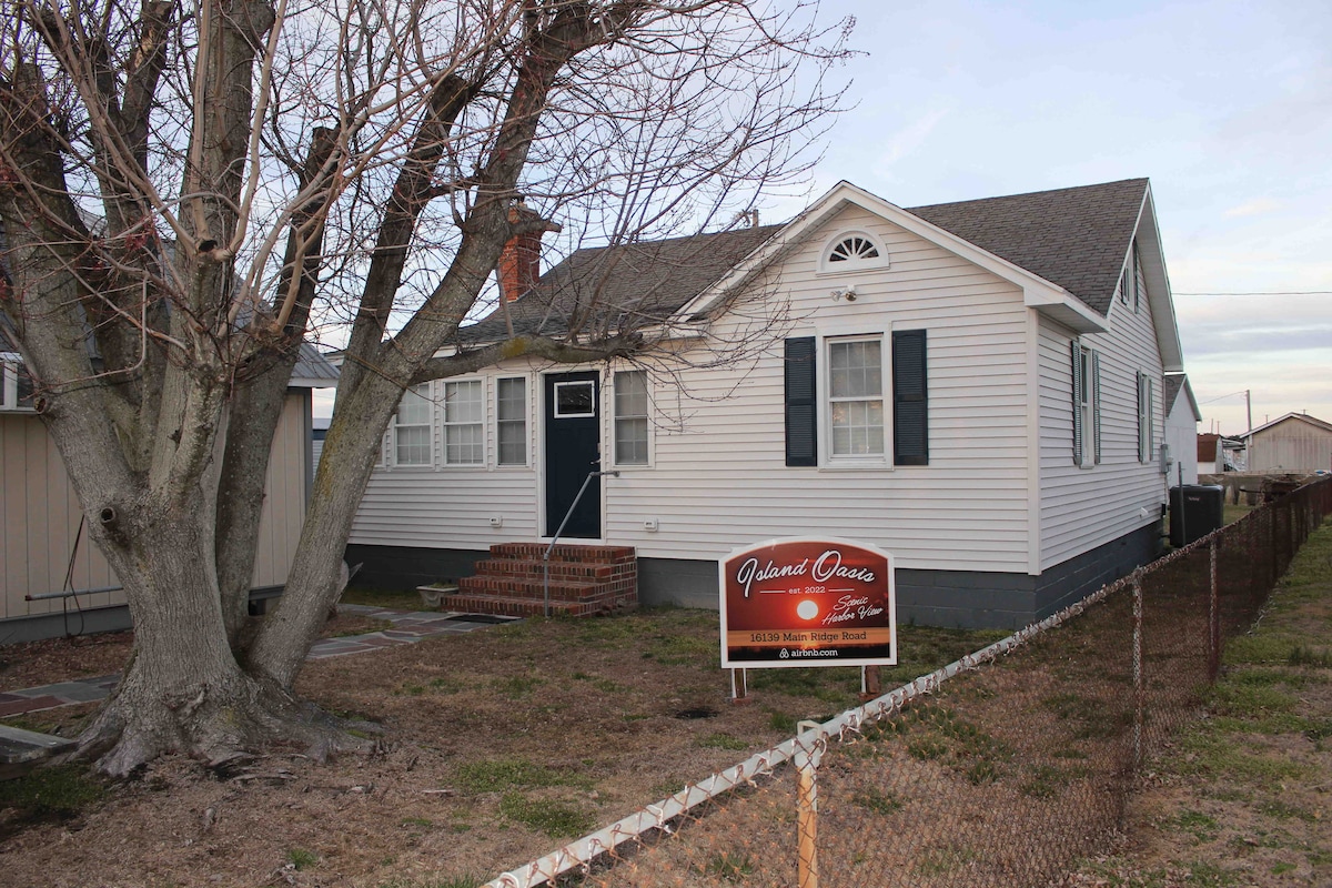 A single-story waterfront home is visible, featuring a light-colored exterior and dark shutters. A welcoming sign is positioned at the front, and a mature tree provides shade in the yard. A simple pathway leads to the entrance, surrounded by a low fence.