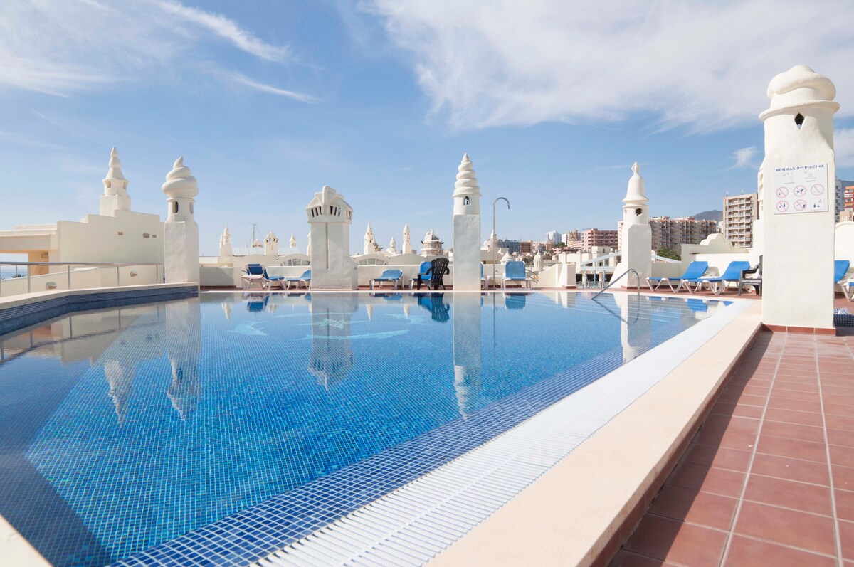 The rooftop pool showcases a spacious area with shimmering blue water and multiple sunbeds arranged around the pool's edge. Imposing white structures provide an elegant backdrop, while clear skies stretch above, enhancing the tranquil ambiance of the space.