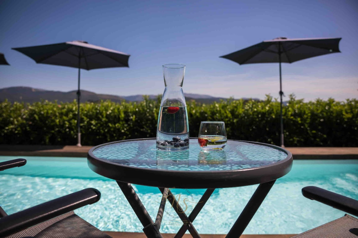 A glass tabletop is set beside a tranquil pool, featuring a pitcher of water and a glass with a cherry. Umbrellas are positioned in the background, providing shade against the clear blue sky and lush greenery surrounding the area.