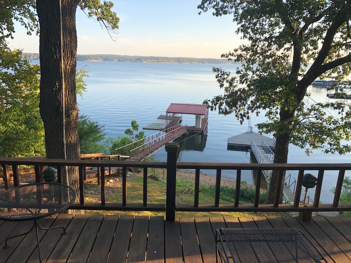 A scenic view is captured from the deck, showcasing a calm lake reflecting the morning light. An elevated walkway leads to a covered dock and two PWC docks, framed by trees on either side. Sturdy railings provide safety along the wooden perimeter.