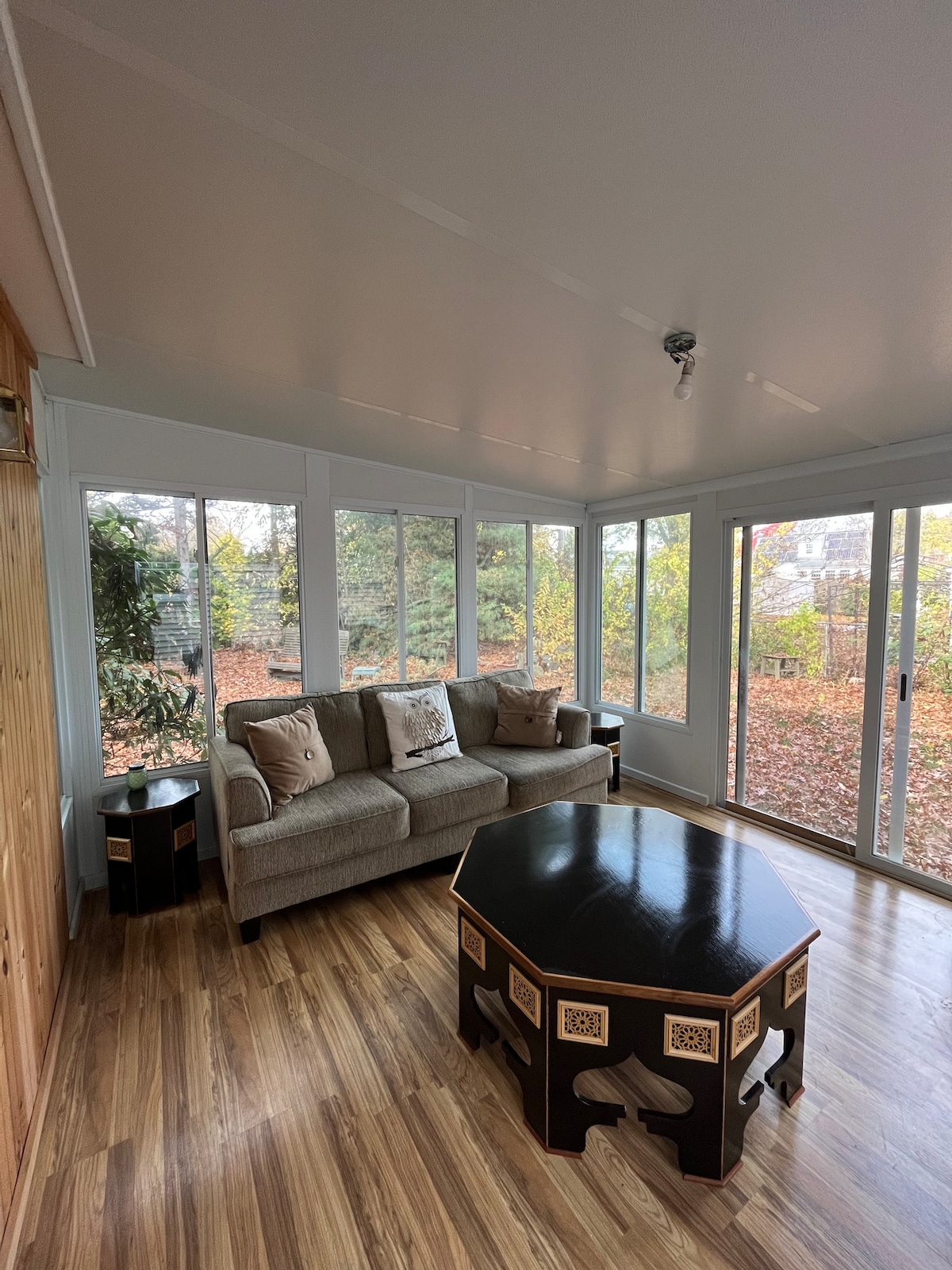 A sunlit sunroom features a comfortable sofa adorned with patterned cushions, situated beside large windows that invite natural light. A dark, intricately designed coffee table occupies the center of the space, with greenery visible outside, enhancing the connection to nature.