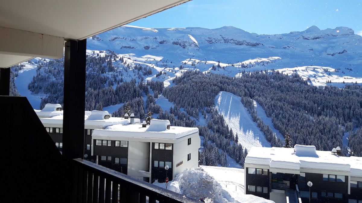 A winter landscape showcases snow-covered mountains and slopes, visible from the balcony. The scene features white snow blanketing the terrain and clusters of trees. In the foreground, modern buildings with dark facades are partially obscured by the railing.