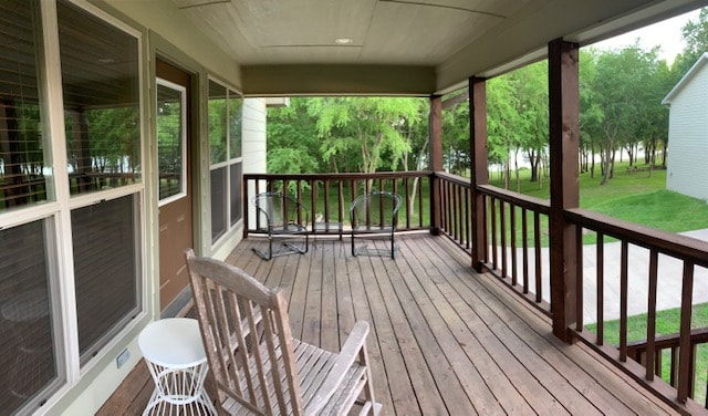 A spacious deck is visible, featuring wooden flooring and a railing made of dark wood. Two chairs are positioned near the edge, offering views of the surrounding trees and the lake in the distance. Natural light filters through the nearby windows.
