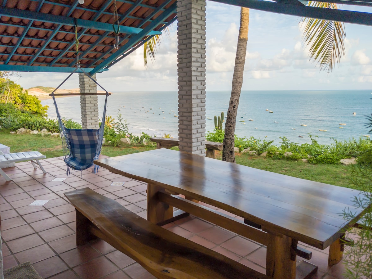 A spacious outdoor area features a large wooden table and a blue and white hammock, set under a tiled roof. Lush greenery frames the view of the sea, dotted with fishing boats. The setting is complemented by palm trees swaying gently in the breeze.