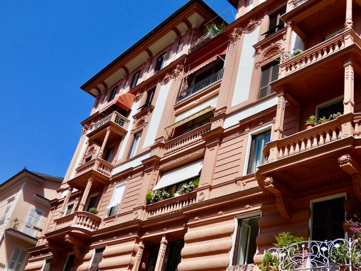 A historic Liberty-style building is depicted under a clear blue sky. Decorative elements and balconies enhance the façade, showcasing ornamental details and lush greenery. The architectural design reflects early 20th-century elegance with a smooth terracotta color.