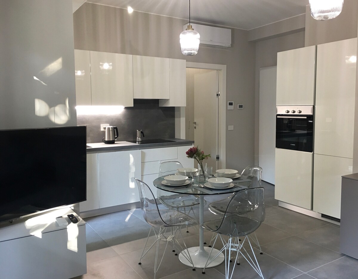 A modern kitchen and dining area are showcased with sleek white cabinetry and gray countertops. A round glass dining table is set for four, with transparent chairs surrounding it. A white refrigerator and built-in oven are visible, along with a coffee maker on the counter.