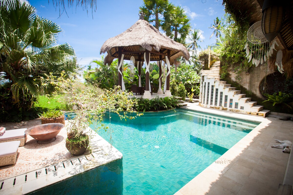 A serene outdoor area features a clear swimming pool surrounded by lush tropical plants. A traditional gazebo is positioned near the pool, providing a shaded seating area. Steps lead from the pool area to a pathway lined with greenery, enhancing the natural atmosphere.