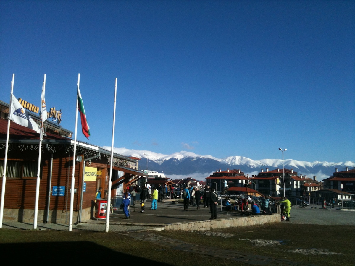 The image features a vibrant ski resort area under a clear blue sky. Snow-capped mountains are visible in the background. People are seen gathered near the resort's entrance, engaging in various activities, with several flags, including the Bulgarian flag, displayed prominently.