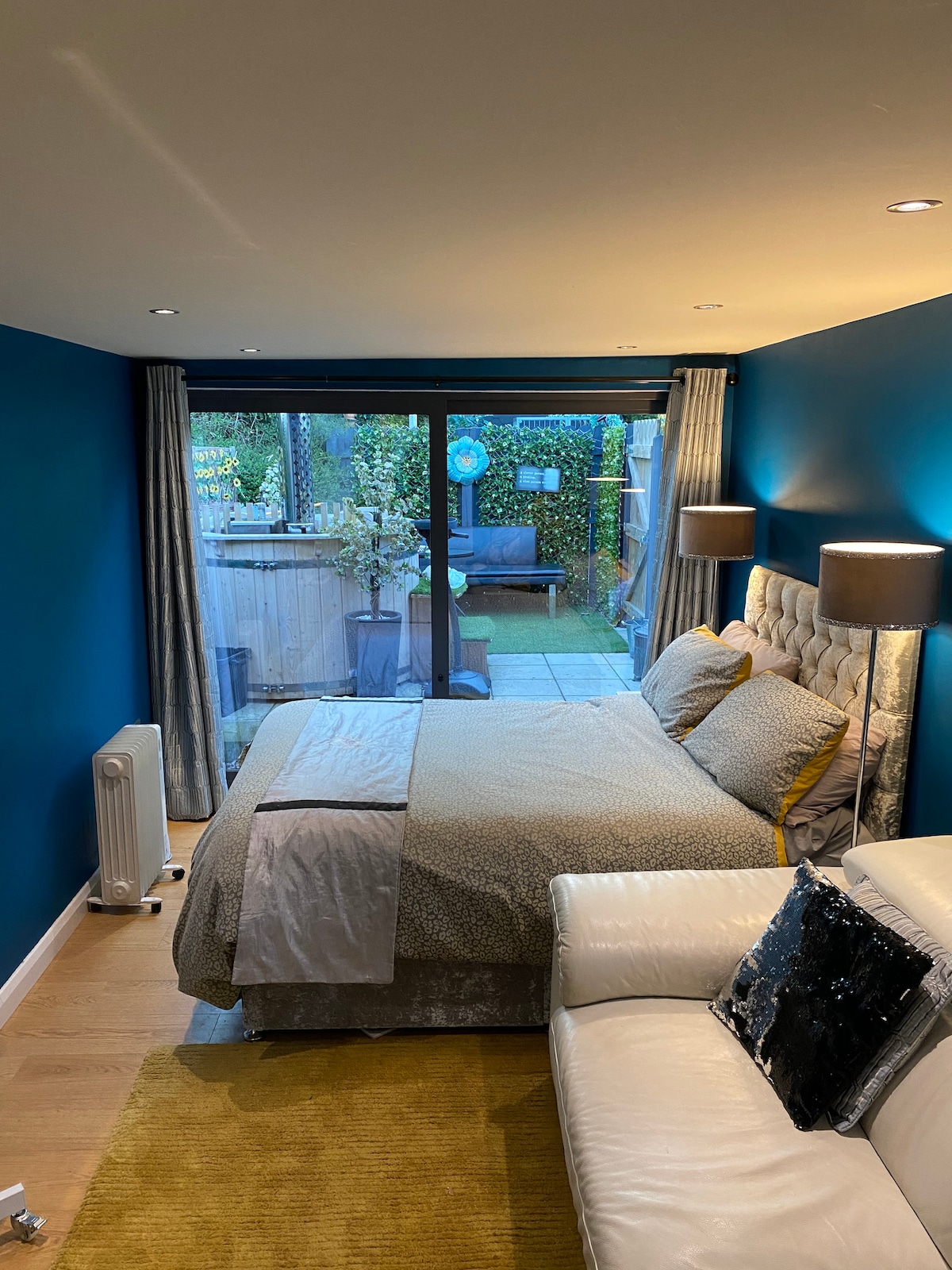 A cozy sleeping area is featured with a spacious bed dressed in neutral-toned bedding. Natural light floods the room through large glass doors leading to a private courtyard. A white sofa with decorative pillows adds comfort, while rich blue walls create a serene atmosphere.