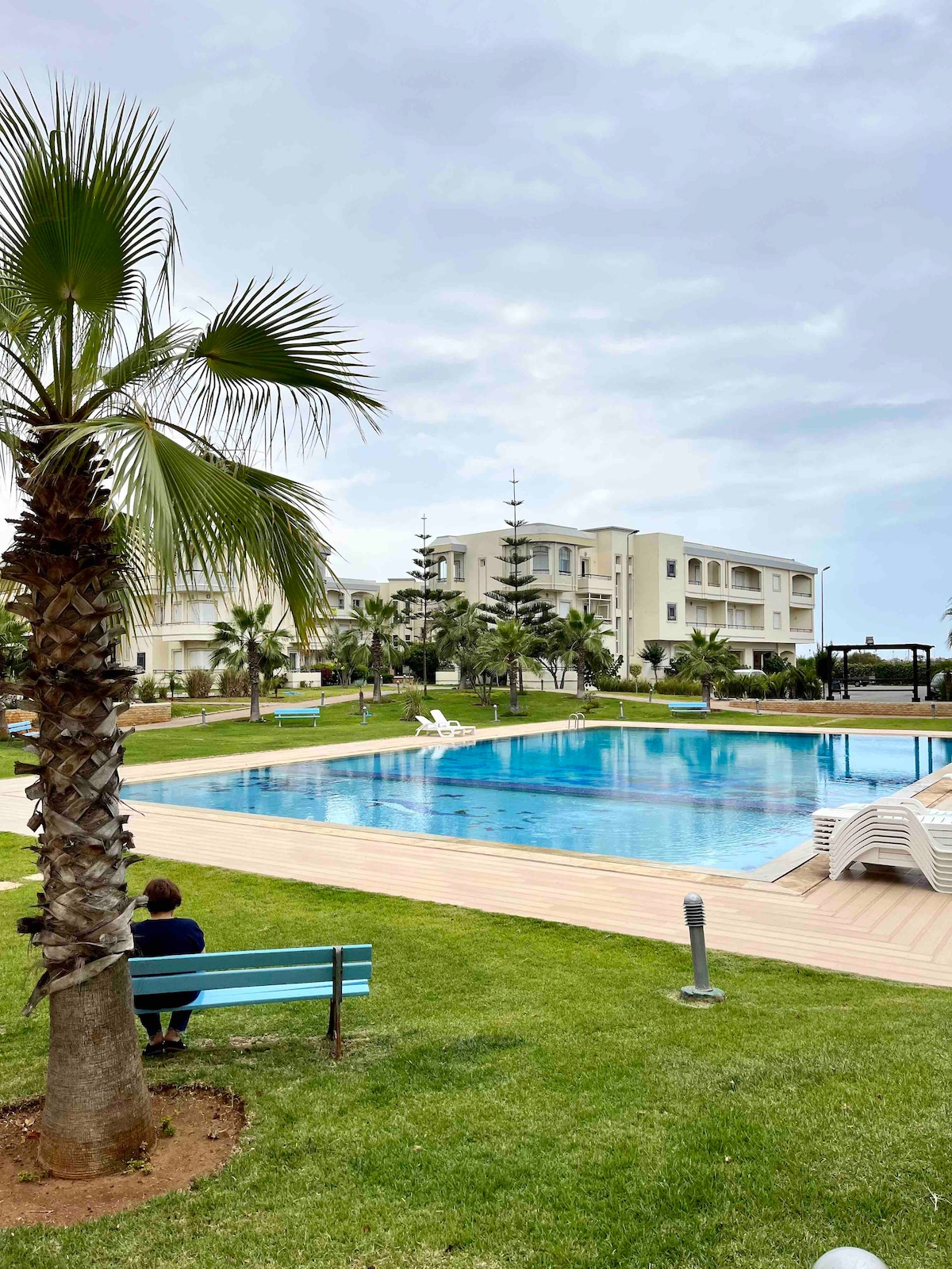 A large swimming pool is surrounded by well-maintained greenery and palm trees. In the foreground, a bench offers a resting spot with a view of the pool and the residential buildings in the background. The atmosphere is peaceful and inviting.