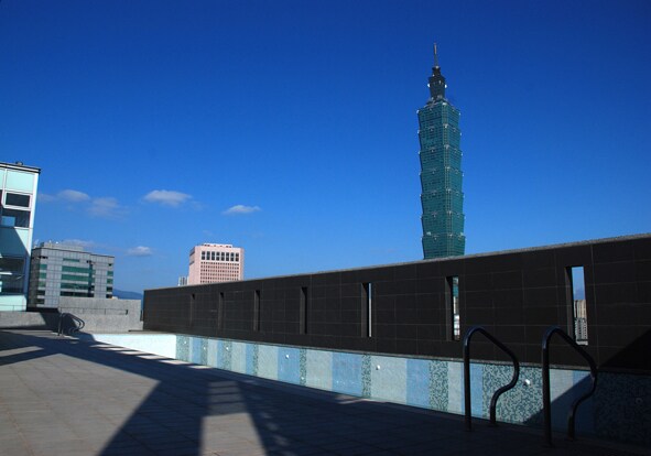 An outdoor swimming pool is visible at the rooftop, framed by a stone wall. The iconic Taipei 101 tower stands tall in the background under a clear blue sky. The pool area is bathed in sunlight, creating a serene atmosphere.
