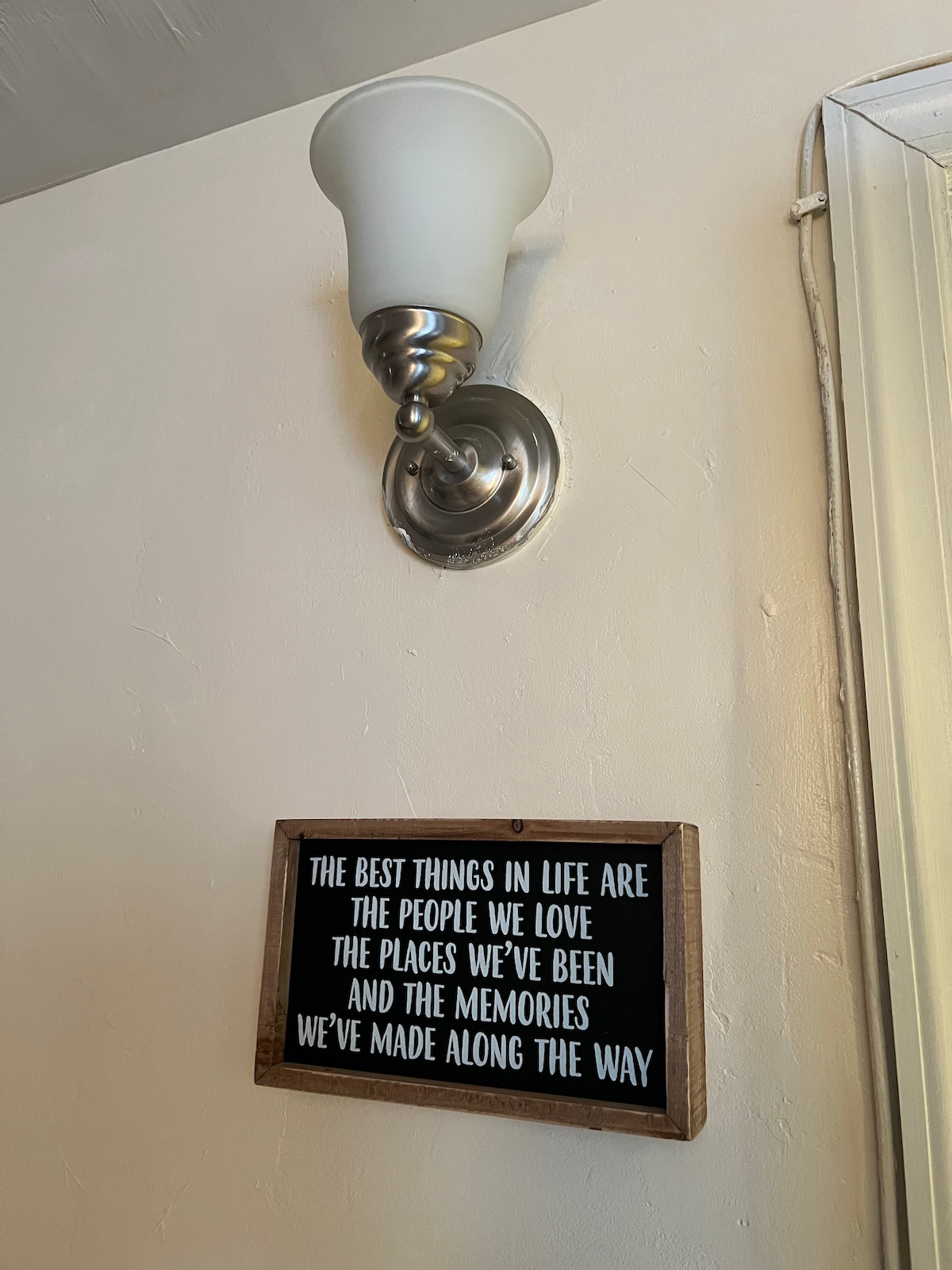 A wall-mounted sconce provides soft lighting above a framed sign. The sign features a wooden border and text that reflects on cherished experiences, emphasizing the value of people, places, and memories.