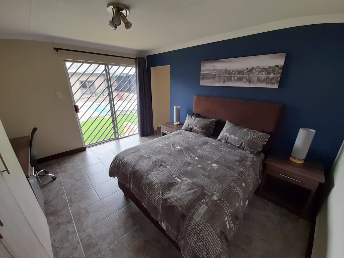 A sleeping area features a spacious bed with patterned bedding, complemented by bedside tables and lamps. Large glass doors provide access to an outdoor pool area, with natural light brightening the room. A simple desk is positioned against one wall.