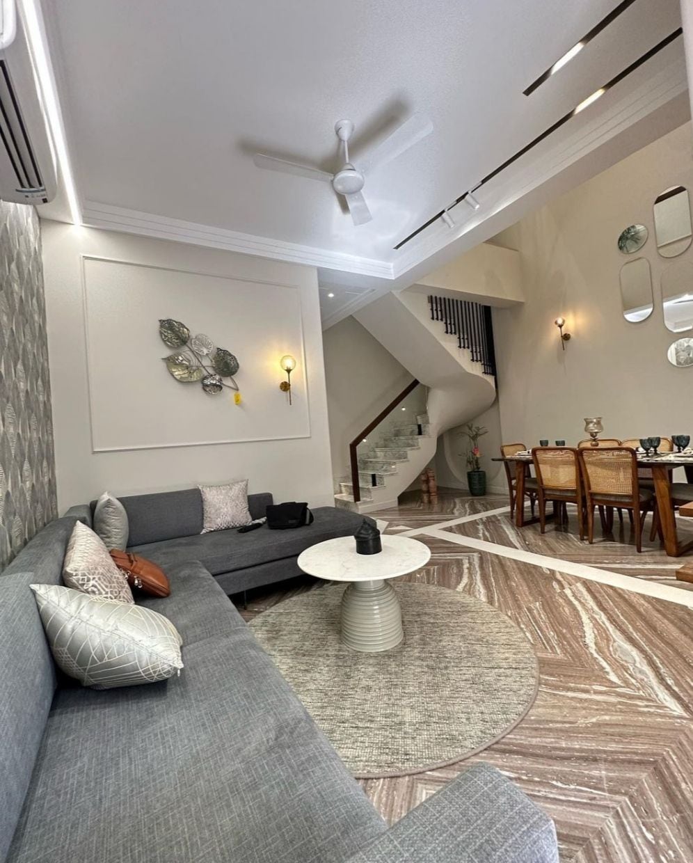 A modern living area features a spacious gray sectional sofa adorned with decorative pillows. A circular coffee table sits atop a round area rug. The dining area is visible in the background, with a wooden table set for several guests. A staircase leads to the upper level.