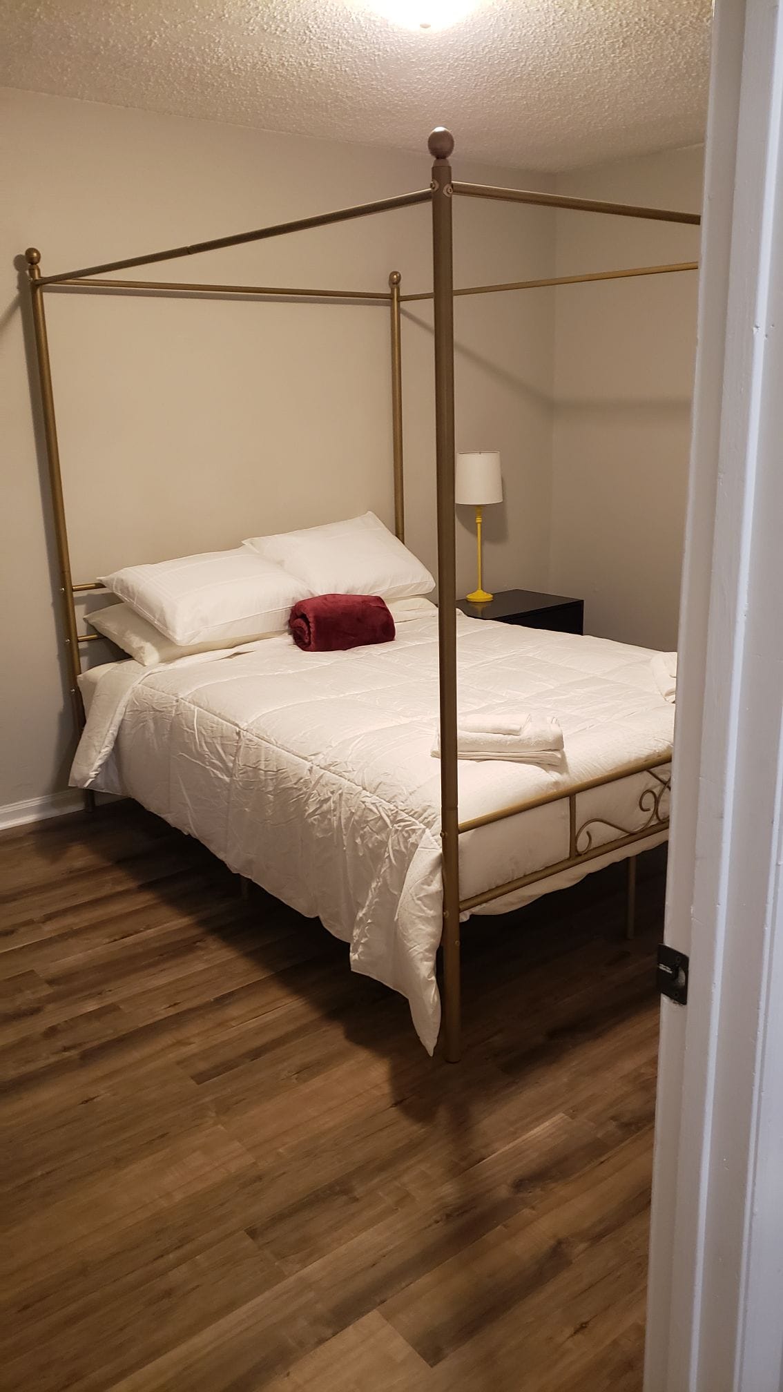 A bedroom is furnished with a gold four-poster bed topped with white linens and pillows. A red towel is neatly placed at the foot of the bed. A yellow lamp stands on a dark nightstand, providing soft lighting in the room.