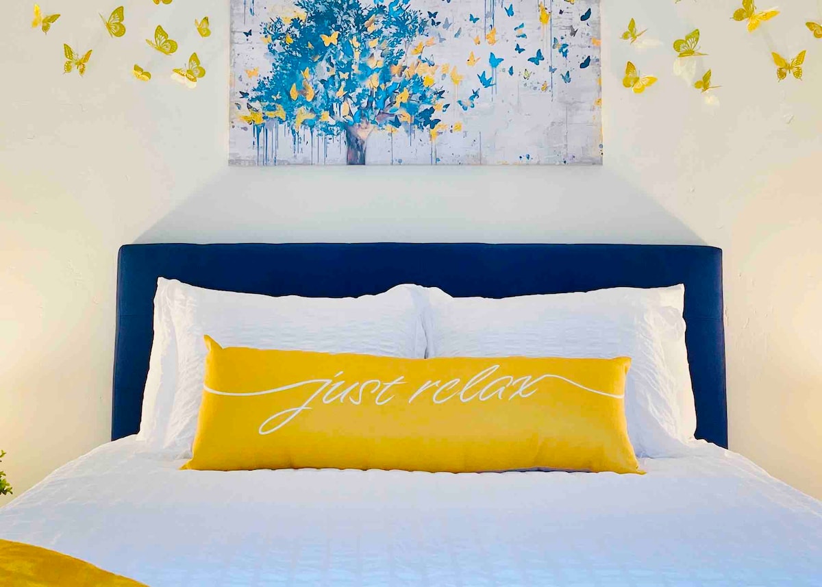 A spacious bed is adorned with crisp white linens and a vibrant yellow accent pillow displaying a calming message. A colorful wall art piece featuring blue flowers and yellow butterflies hangs above, creating a cheerful atmosphere. Complementary butterfly decorations are scattered artistically on the wall.