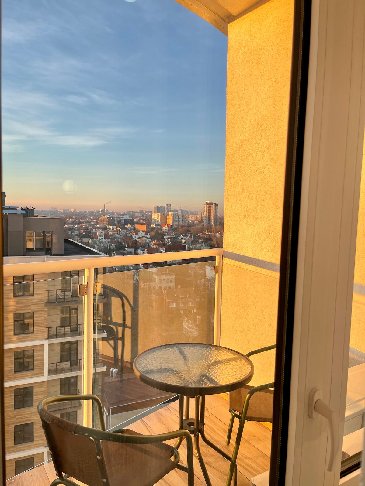 A balcony view is captured, featuring a small round table and two chairs. The space overlooks a city skyline bathed in warm evening light. Tall buildings and distant views of the horizon are visible, creating an inviting outdoor seating area.