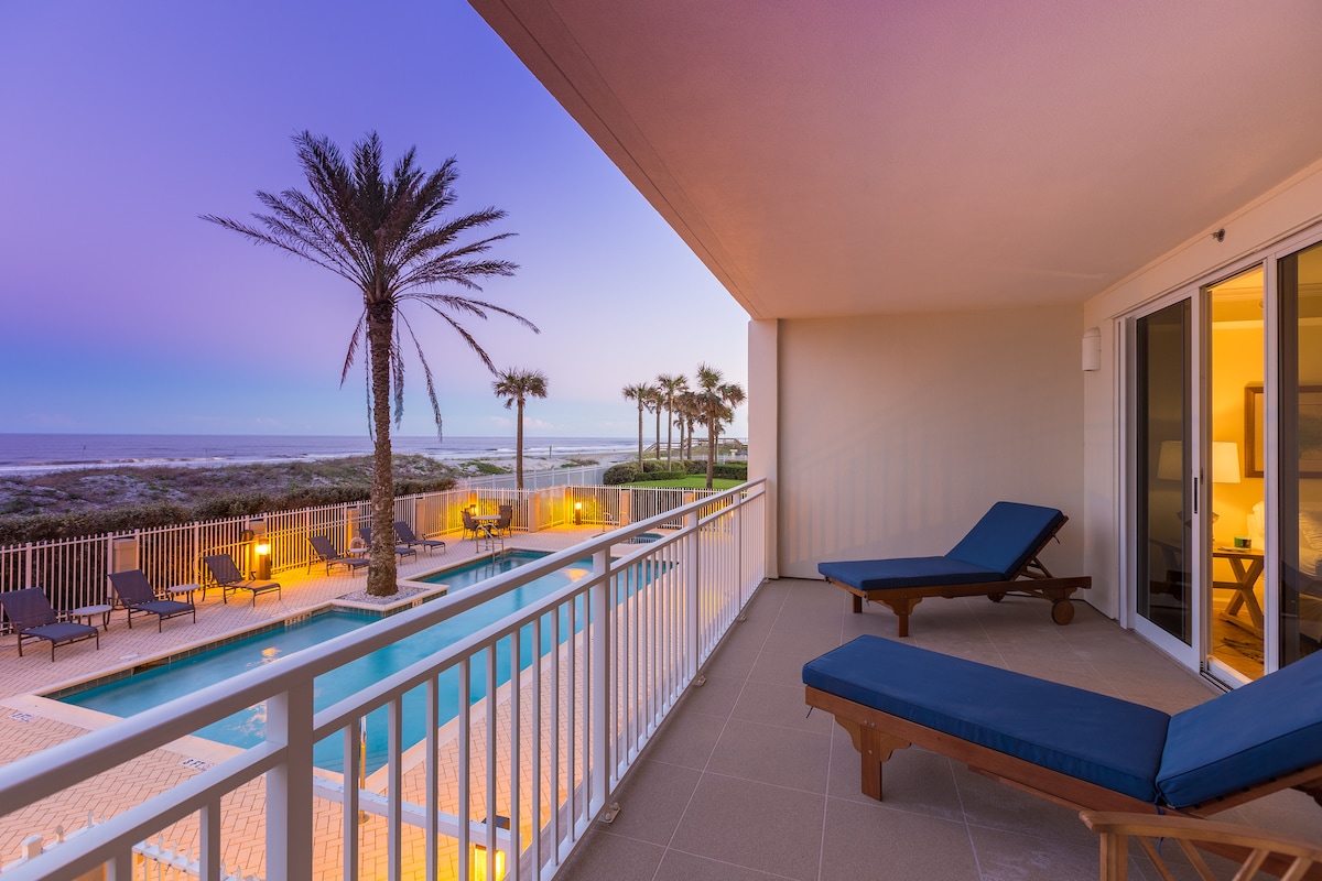 An over-sized balcony overlooks the Atlantic Ocean, featuring two lounge chairs positioned for relaxation. The view includes palm trees and a swimming pool below, with a gentle sunset lighting the sky in soft purple hues.