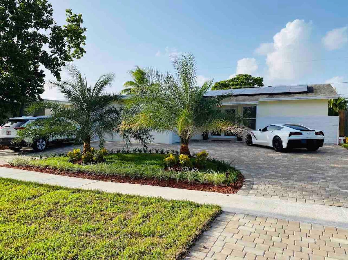 The exterior of a single-story home is showcased, featuring a paved driveway with two vehicles parked. Lush palm trees and vibrant landscaping enhance the curb appeal. A bright blue sky is visible above, creating a tranquil atmosphere.