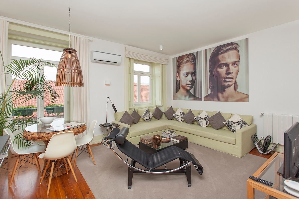 The living area features a light green sectional sofa adorned with patterned cushions and an artistic wall display. A round dining table with white chairs is visible, along with a floor lamp and a television. Natural light enters through large windows, creating a relaxing space.