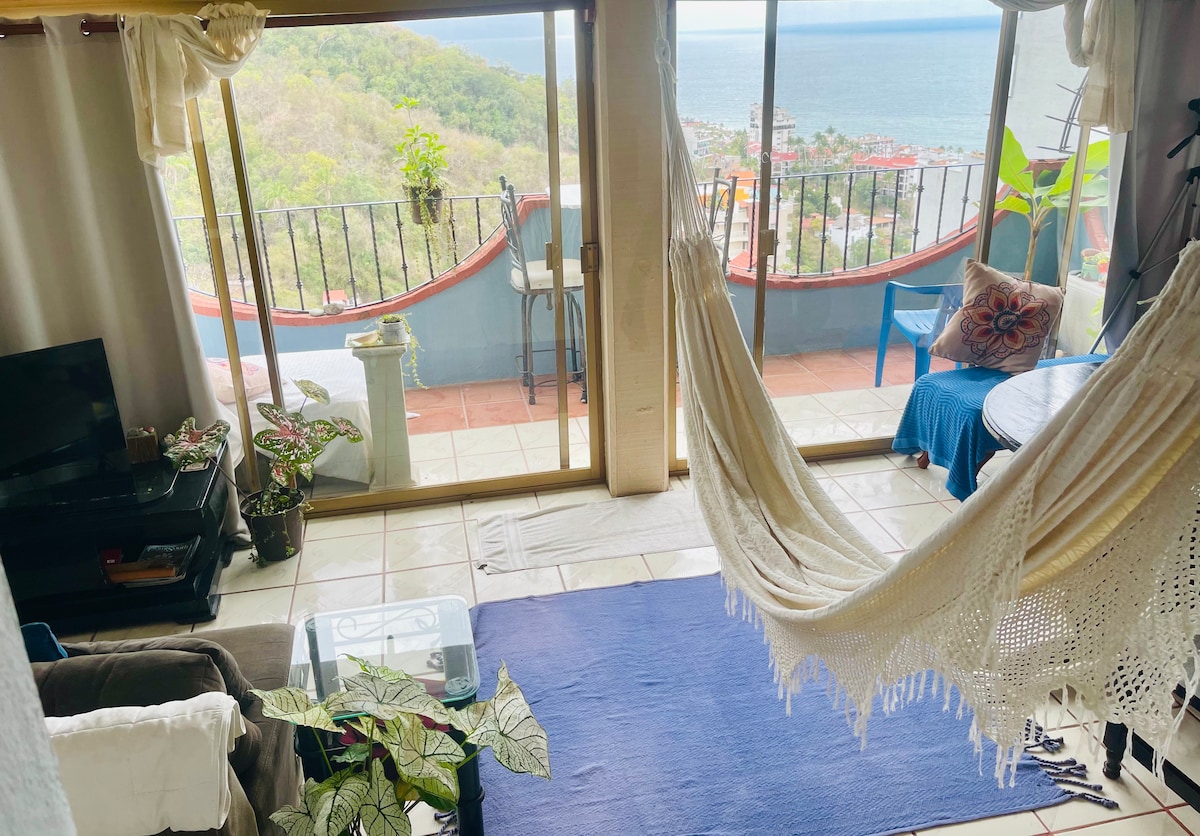 A cozy living space features large sliding glass doors, providing access to a terrace with ocean views. A hammock drapes between the walls, and potted plants add greenery. Light-colored curtains softly filter sunlight, enhancing the warm atmosphere of the room.