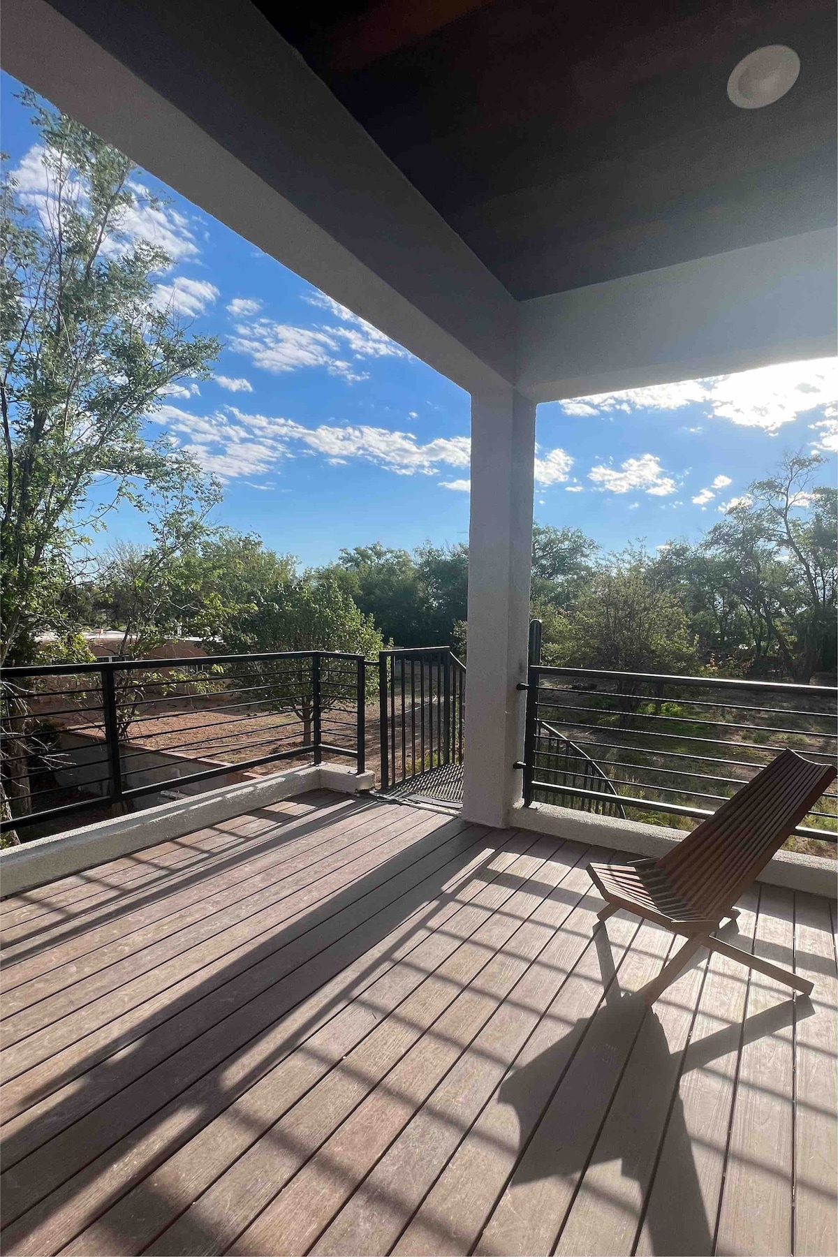 A spacious patio area features wooden flooring and a single outdoor chair. Sunlight filters through the railing, creating a pattern on the ground. Surrounding greenery and blue sky contribute to a bright and open atmosphere.