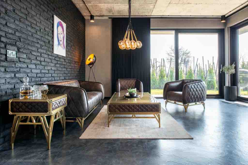 A modern living area features dark brick walls and large windows allowing natural light to illuminate the space. Comfortable leather sofas with a geometric design are arranged around a stylish coffee table. A piece of artwork adds a splash of color, enhancing the room's aesthetic.