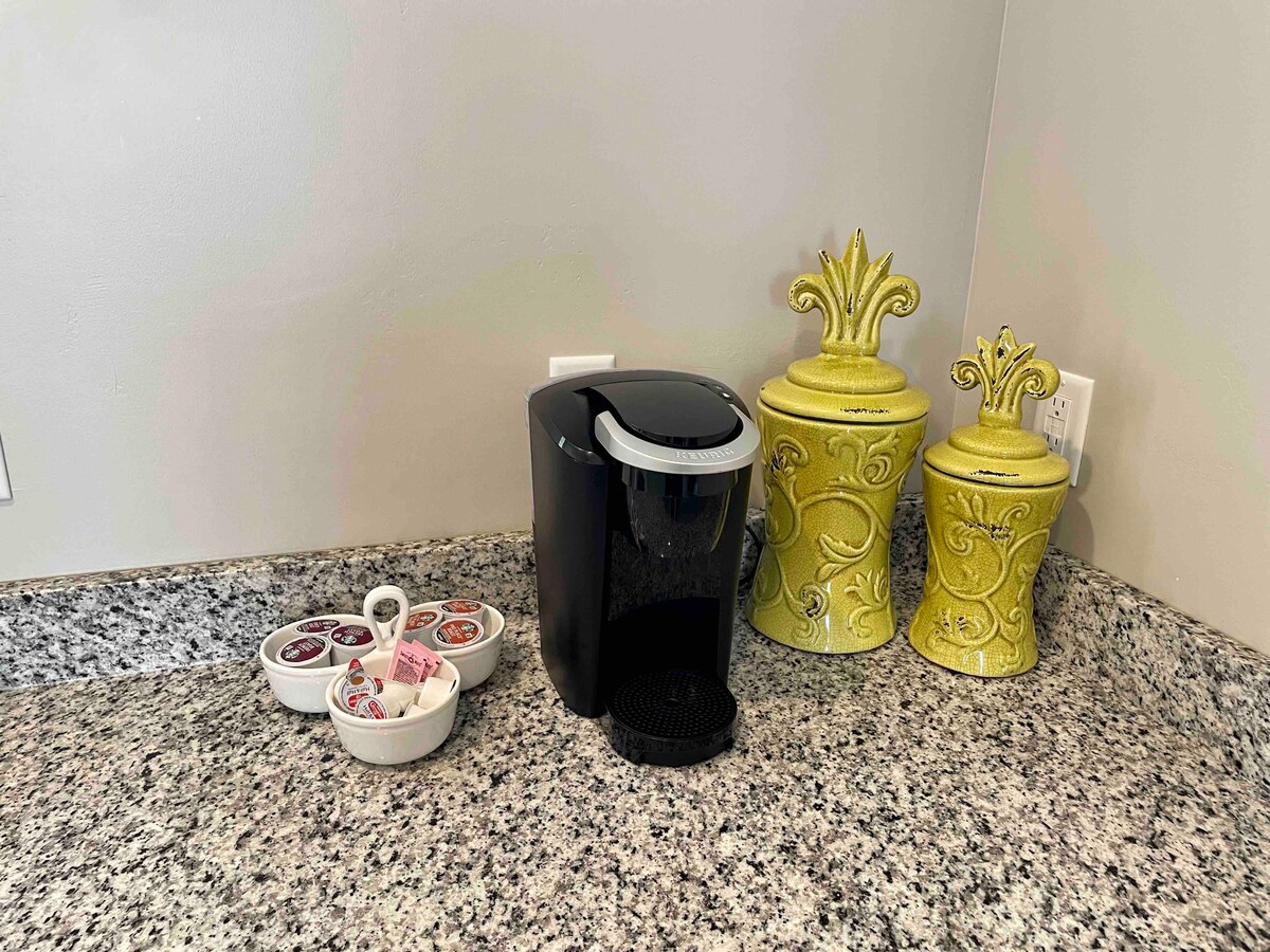 A sleek black coffee maker is positioned on a speckled granite countertop. Next to it, decorative yellow canisters add a bright touch to the space. A small dish of coffee pods and creamers is also visible, offering convenience for guests.