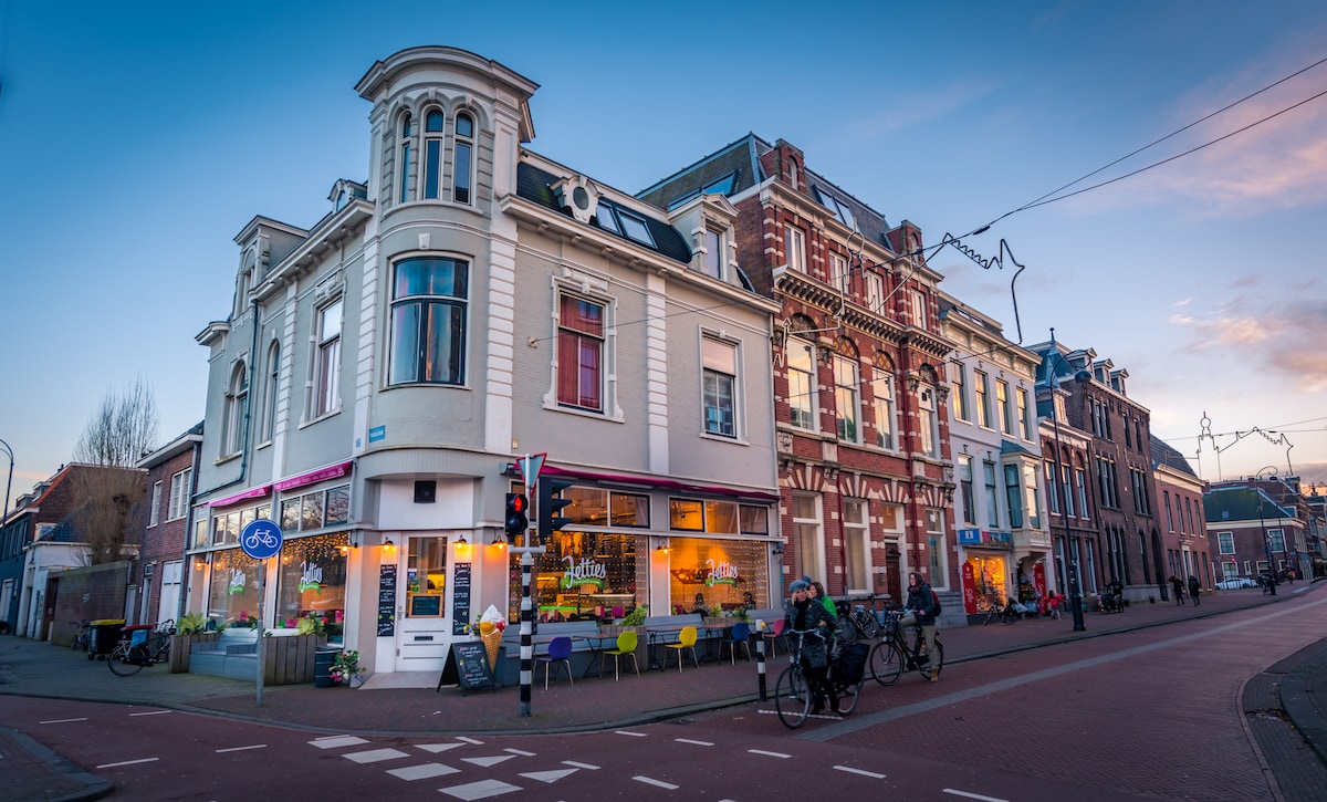 A charming building stands at the intersection, showcasing a blend of architectural styles. The ground floor features large windows with colorful outdoor seating, while bicycles are parked nearby. Streetlights and vibrant storefronts contribute to the lively atmosphere of this Haarlem neighborhood.