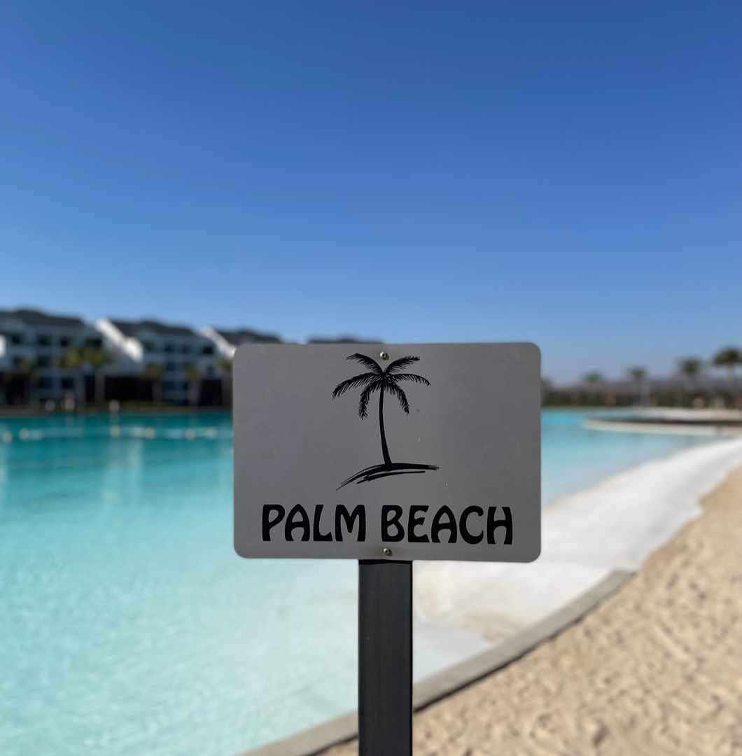 A sign displaying the text 'PALM BEACH' is positioned beside a calm pool, with a palm tree graphic. The surrounding area features sandy edges, while a row of structures is visible in the background under a clear blue sky.