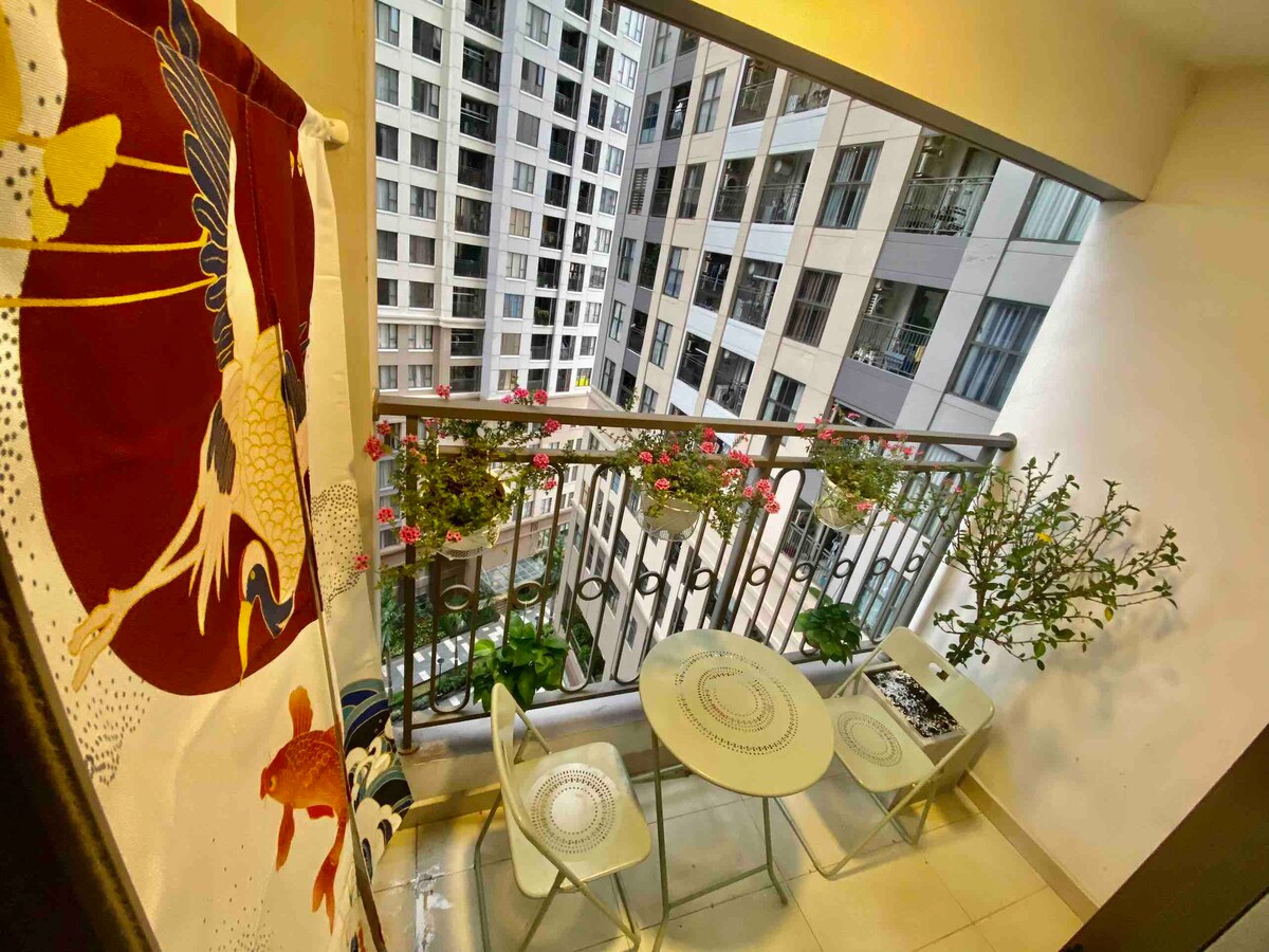 A small balcony features a round table and two chairs, surrounded by decorative plants. A colorful curtain with a bird motif provides privacy and adds visual interest. Buildings are visible in the background, creating an urban atmosphere.