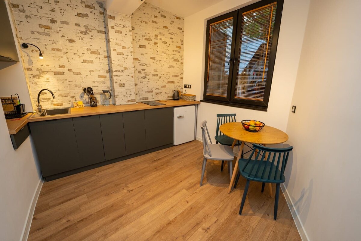 A modern kitchen area features a light wood countertop and dark cabinetry against a textured brick wall. A round wooden table with blue and gray chairs is positioned near a window, allowing natural light to illuminate the space.