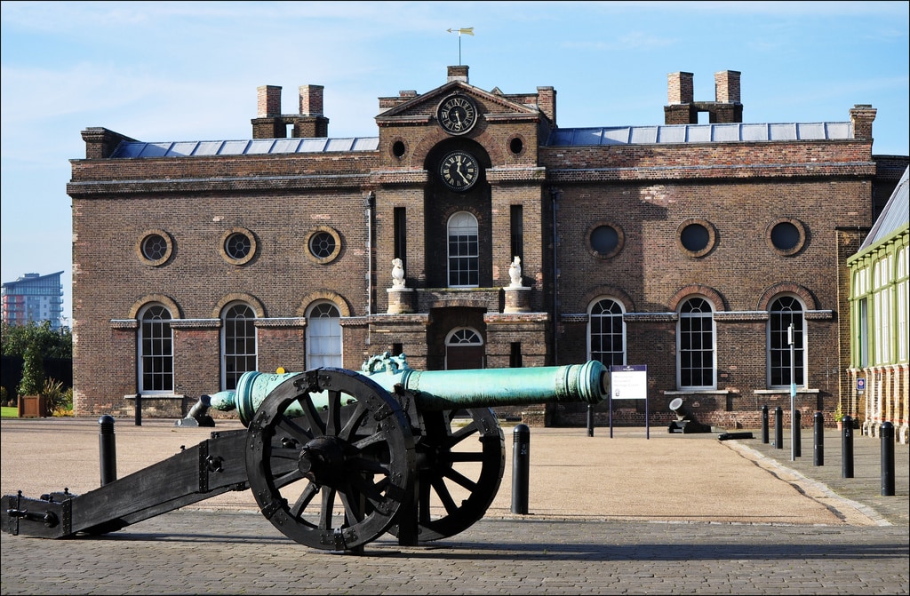A historic brick building features large round windows and a prominent clock above the entrance. In the foreground, a vintage cannon is positioned on a paved area, highlighting the architectural details of the structure, which is accented by a vibrant green roof.