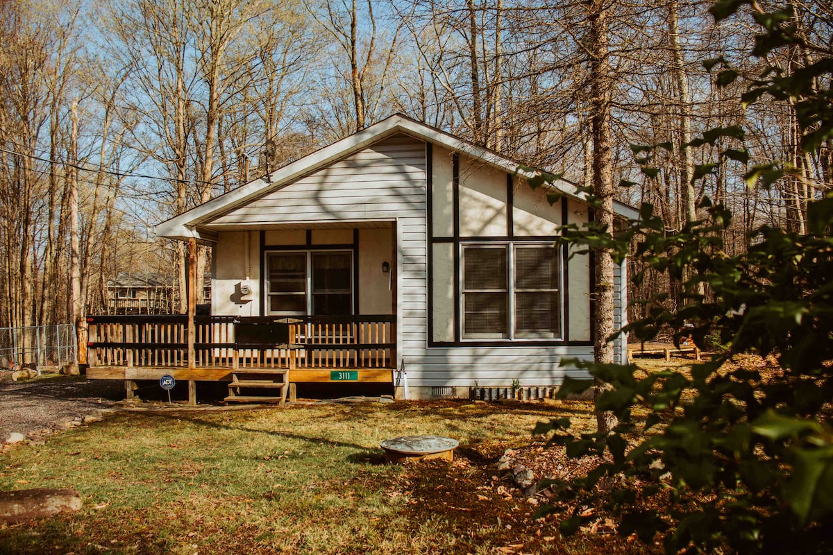 The exterior of a charming single-story cottage is presented, featuring a covered front porch with wooden railing. Large windows allow natural light inside. Surrounding trees provide a tranquil backdrop, while a grassy area leads to a gravel driveway.